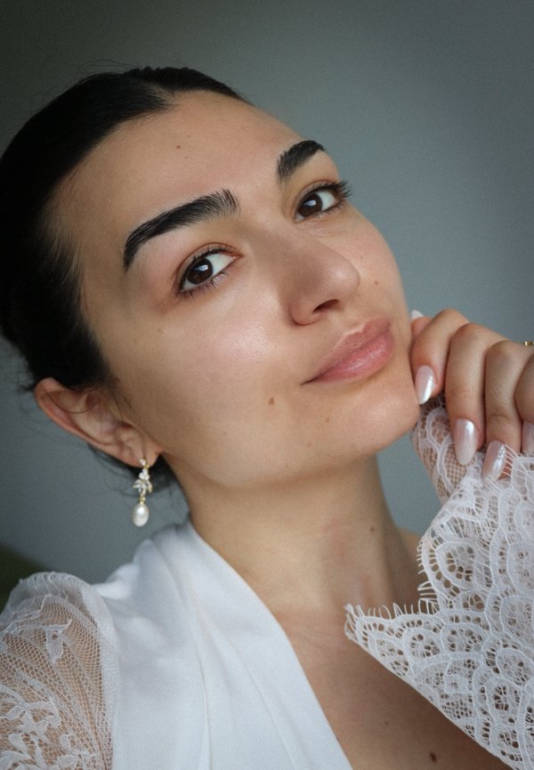 Jeune femme aux cheveux foncés portant des boucles d'oreilles en perles et un vêtement en dentelle blanche, touchant doucement son visage avec des ongles manucurés.