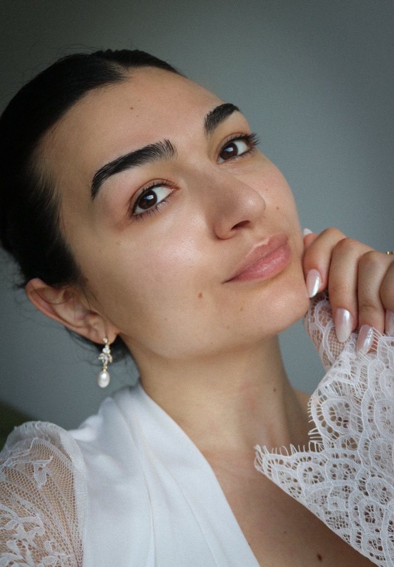 Jeune femme aux cheveux foncés portant des boucles d'oreilles en perles et un vêtement en dentelle blanche, touchant doucement son visage avec des ongles manucurés.