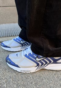 Λευκά και ναυτικά μπλε αθλητικά παπούτσια Nike με πλέγμα, εμφανή μαύρη φτέρνα και υφασμάτινη σόλα. Φορεμένα με μαύρα παντελόνια από χοντρό βελούδο.