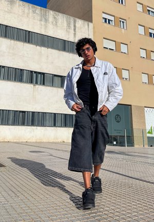 Chaqueta de denim blanca sobre una camiseta negra sin mangas, combinada con culottes negros sueltos y zapatillas negras; usando gafas de sol y auriculares. Entorno urbano.