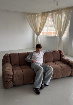 Sofá marrón de felpa con diseño suelto y capitoné y brazos redondeados. Paredes gris claro y cortinas transparentes en una habitación espaciosa y minimalista.