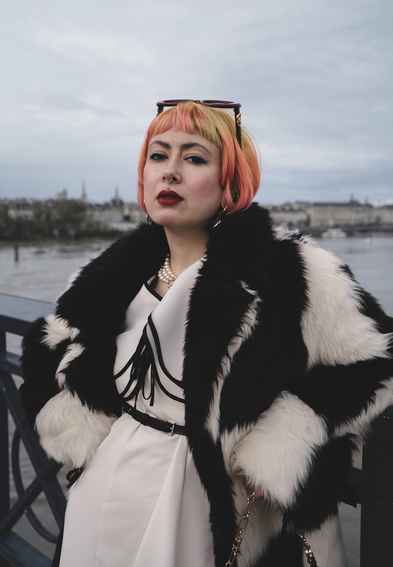 Cappotto in pelliccia ecologica bianco e nero sopra un vestito crema con accenti neri, con collana di perle e occhiali da sole oversize.