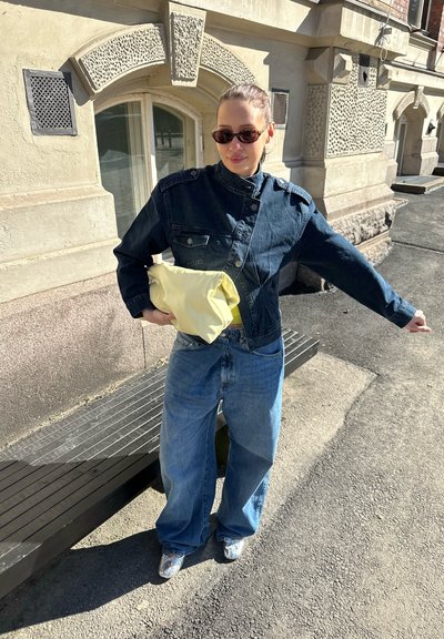Mujer con chaqueta vaquera oscura, vaqueros azules sueltos, gafas de sol, sosteniendo una bolsa amarilla, de pie en una acera cerca de un edificio de piedra con textura.