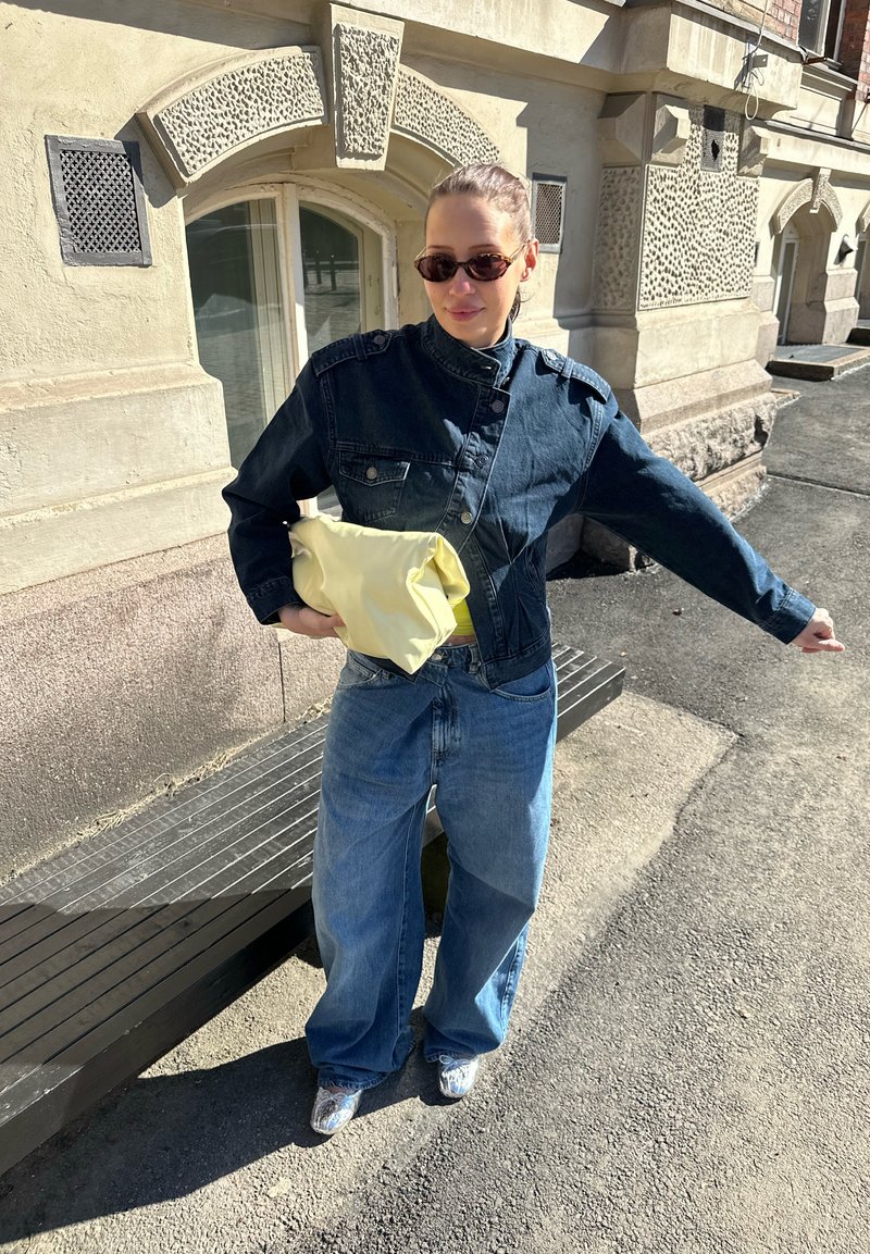 Femme portant une veste en jean foncée, un jean bleu ample, des lunettes de soleil, tenant un sac jaune, debout sur un trottoir près d’un bâtiment en pierre texturée.