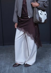 Persoon in wijde witte broek, bruin top met kant en asymmetrische snit, grijze jas, die een grote bruine handtas met witte vlekken vasthoudt, staande op een betonnen vloer.
