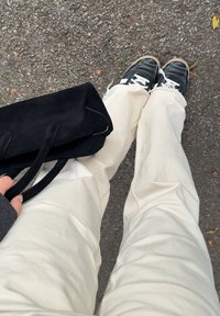 Personne portant un pantalon beige et des baskets noires debout sur l'asphalte, tenant un sac à main noir avec deux poignées.