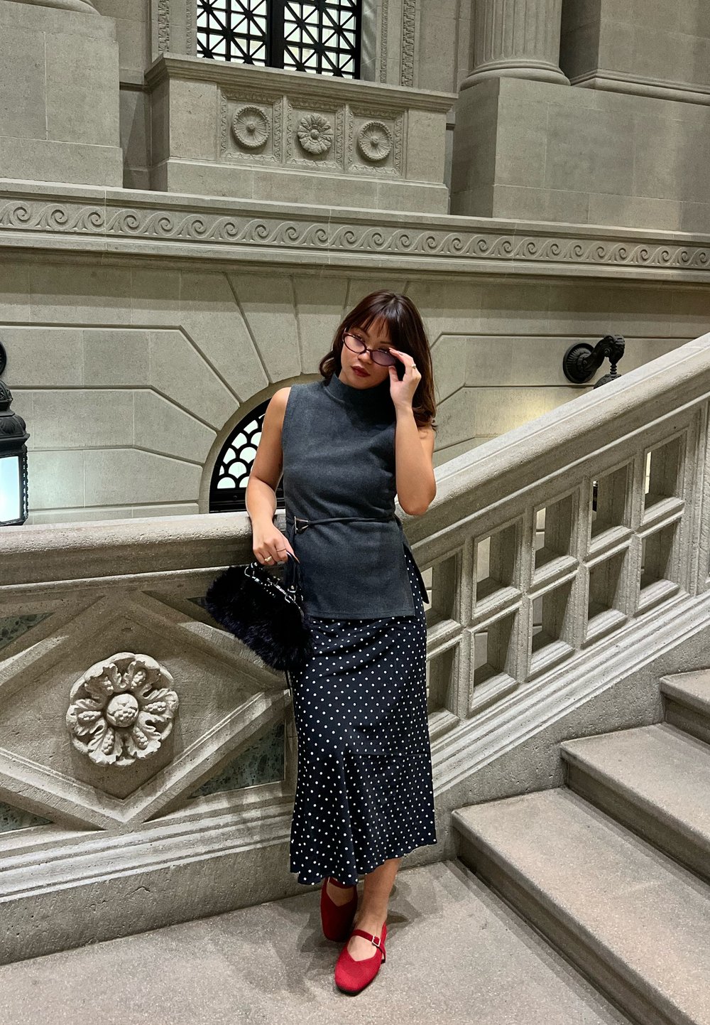 Femme ajustant ses lunettes, portant un haut gris sans manches, une jupe noire à pois, des chaussures rouges, tenant un sac à main en fourrure noire, debout près d'un escalier en pierre.