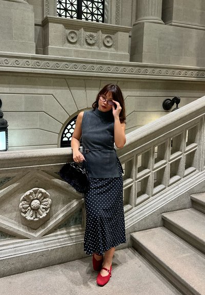 Mujer ajustándose las gafas, lleva una blusa gris sin mangas, falda negra con puntos blancos, zapatos rojos, sostiene un bolso de piel negro y está de pie junto a una escalera de piedra.