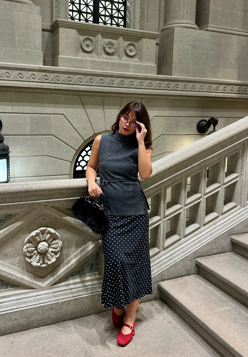 Vrouw die haar bril rechtzet, draagt een mouwloos grijs topje, een zwarte rok met witte stippen, rode schoenen, en houdt een zwarte bont handtas vast, staand bij een stenen trap.