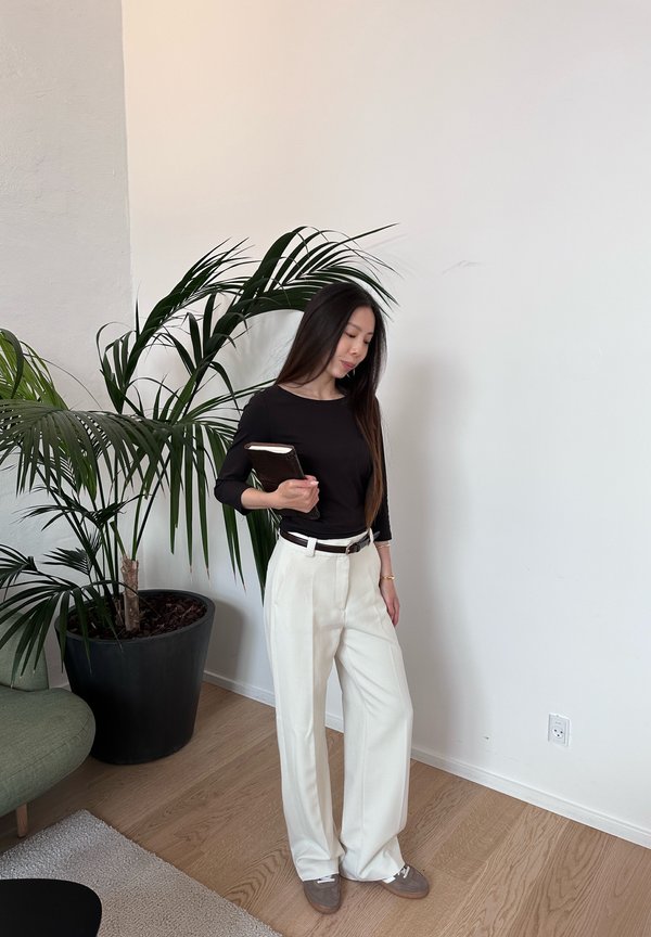 Una mujer con una camiseta negra y pantalones blancos sostiene un libro oscuro junto a una palmera grande en una maceta, en una habitación luminosa.