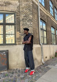 Μαύρο άσπρο χωρίς μανίκια πουκάμισο, χαλαρό σκούρο τζιν παντελόνι, κόκκινες αθλητικές σαμπό και μαύρη μπερέτα. Σκληρός φτιαγμένος με πέτρες δρόμος και τοίχος από τούβλα στο βάθος.