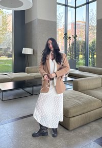 Καφέ σακάκι πάνω από ένα λευκό δαντελένιο φόρεμα, συνδυασμένο με μαύρες γόβες. Γυαλιά ηλίου στο χέρι. Σύγχρονος εσωτερικός χώρος με μεγάλες παράθυρες και πράσινες θέες.