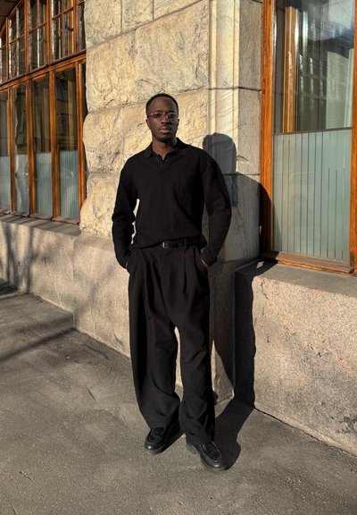 Hombre con camisa negra, pantalones holgados negros y gafas, está de pie con las manos en los bolsillos frente a una pared de piedra con ventanas con marcos de madera a la luz del sol.