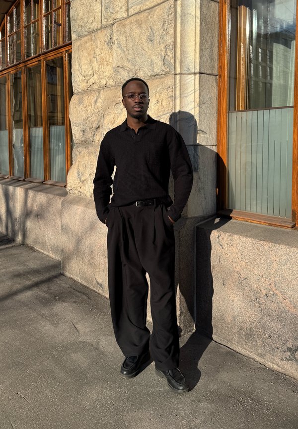 Muž v čiernej košeli, voľných čiernych nohaviciach a okuliaroch stojí s rukami v kapsách pri kamennej stene s drevenými rámy okien na slnku.