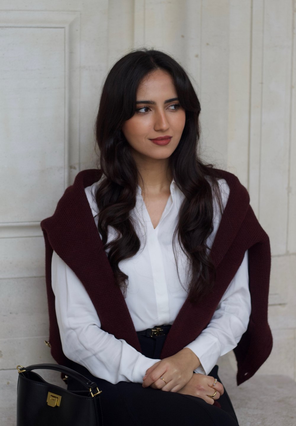 Maroon sweater draped over a white blouse, black skirt, and a black handbag with gold accents. Smooth texture and tailored fit visible.