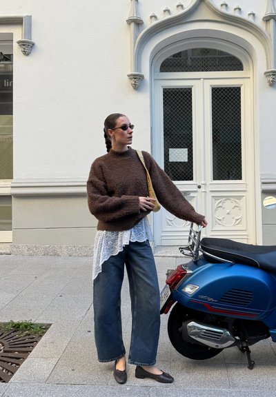 Suéter marrón peludo con mangas sueltas, superpuesto a una blusa blanca con encaje en los bordes. Jeans anchos azules y zapatos planos oscuros. Scooters azules al lado.