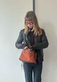 Oranža auduma rokassoma ar strukturētu formu un metāla akcentiem. Iespējas īsas rokturi un brūna apdare, ko tur cilvēks melnā žaketē.