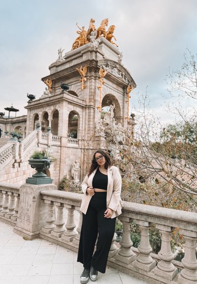Estructura de piedra con acentos dorados, arcos ornamentados y estatuas decorativas. En primer plano, hay una mujer con un blazer claro y un atuendo negro.