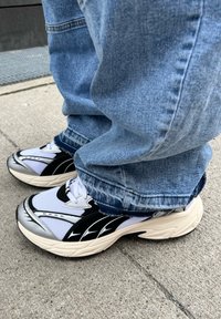 Sneakers bianchi e neri con una tomaia in mesh testurizzato, dettagli argento e una suola spessa, indossati con jeans in denim blu a vestibilità ampia.