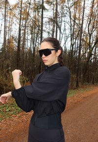 Giacca sportiva nera con maniche arricciate, chiusura con cerniera e vita aderente; abbinata a occhiali da sole neri oversize, ambientata in un'area boschiva.