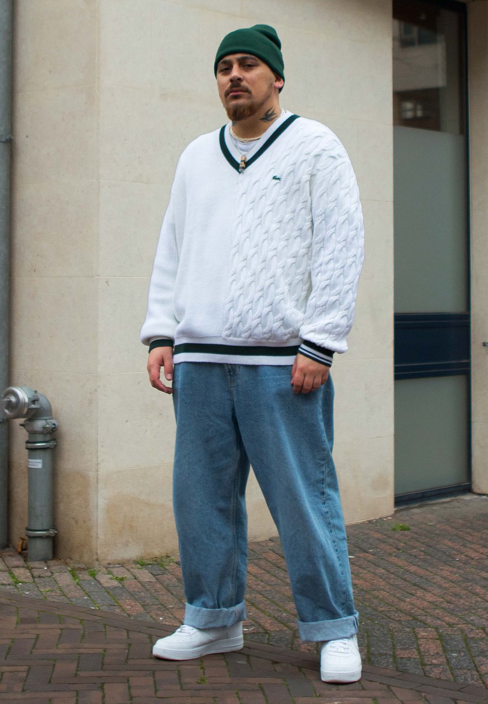 Fehér kötött pulóver zöld részletekkel, világoskék bő farmerral és fehér sportcipővel p�árosítva. Sötétzöld sapka a fején.