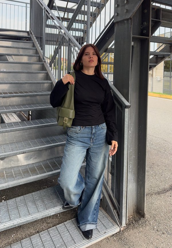 Mujer con cabello oscuro que lleva una camiseta negra y vaqueros anchos está de pie en unas escaleras metálicas exteriores sosteniendo una bolsa verde sobre su hombro.