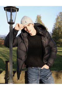 Melna polsterēta jaka, melns rakstains džemperis, gaismas pelēka cepure, stāvot pie laternas zaļā āra vidē.