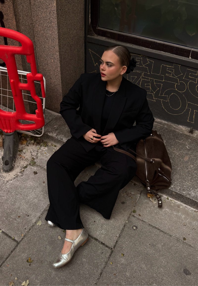 Černý oversized blazer a kalhoty, v kombinaci se stříbrnými Mary Jane botami. Hnědá kožená taška leží poblíž na texturovaném pozadí.