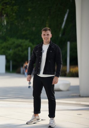 Chaqueta de denim negra sobre una camiseta blanca con texto, pantalones negros arremangados y zapatillas grises. Sosteniendo gafas de sol en una mano.