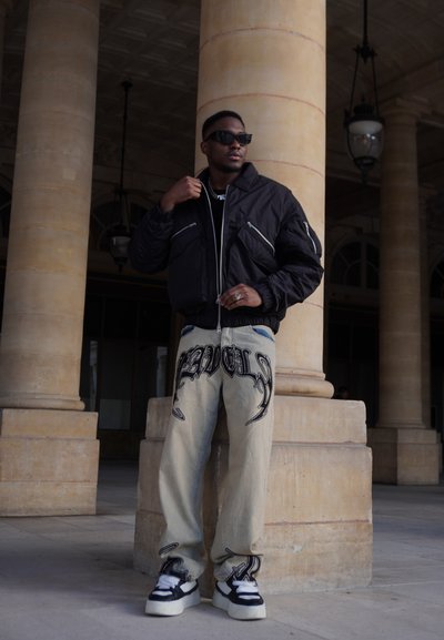 Cazadora bomber negra con bolsillos con cremallera, camiseta negra, jeans de denim claros con texto gráfico negro, y zapatillas en blanco y negro.