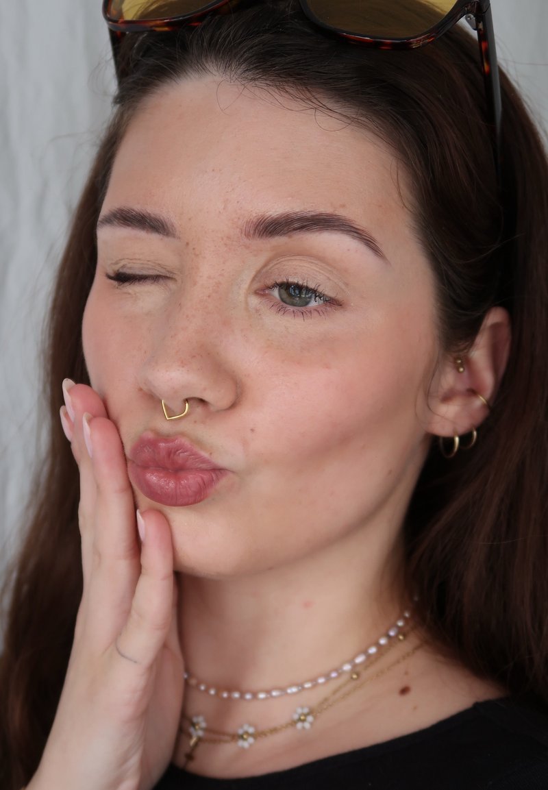 Jonge vrouw met zonnebril op haar hoofd, knipogend, lippen tuitend, wang aanrakend, met een neusring, oorbellen en gelaagde kettingen met bloemenhangers.
