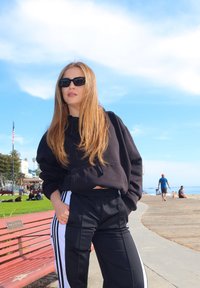 Crni izrezani dukserica s dugim rukavima i crne hlače s bijelim prugama uz strane. Pozadina prikazuje plažu i šetnicu.