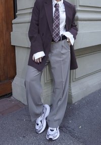 Osoba opierająca się o ścianę, ubrana w fioletową marynarkę w prążki, kraciasty krawat, szare spodnie i białe buty sportowe.