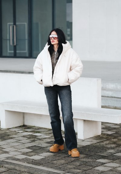 Chaqueta blanca abullonada con capucha, camiseta interior con estampado, vaqueros oscuros y zapatos plataforma amarillos. Las gafas de sol reflejan la luz. Fondo de concreto.