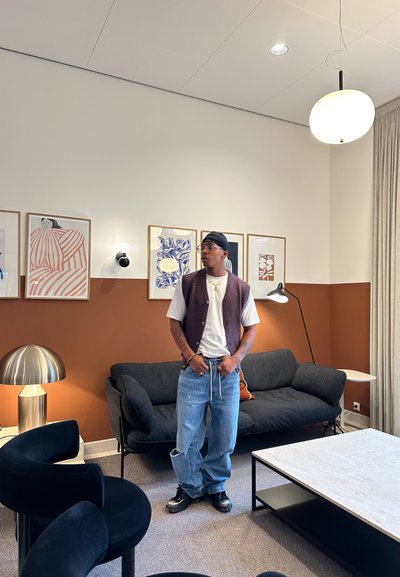 Hombre con una camiseta blanca, chaleco morado, jeans anchos y gafas, de pie en una sala de estar moderna con obras de arte enmarcadas, un sofá y lámparas.