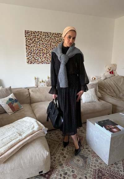 Mujer con vestido negro de lunares, bufanda gris, pañuelo beige en la cabeza y tacones negros está en la sala de estar sosteniendo un bolso negro.