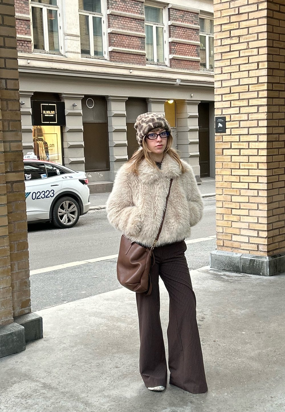 Ung kvinde iført en beige jakke i kunstpels, leopardmønstret hat, brune bukser og bærende en brun crossbody-taske på en byfortov.
