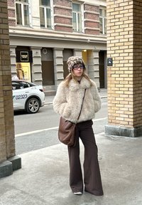 Ung kvinde iført en beige jakke i kunstpels, leopardmønstret hat, brune bukser og bærende en brun crossbody-taske på en byfortov.