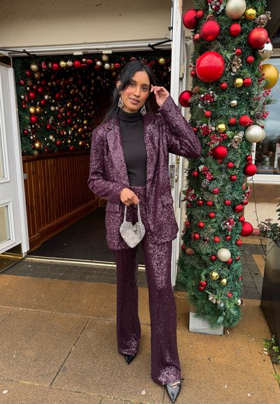 Traje de lentejuelas morado con un blazer ajustado y pantalones acampanados, combinado con un cuello de tortuga negro y un bolso en forma de corazón plateado.