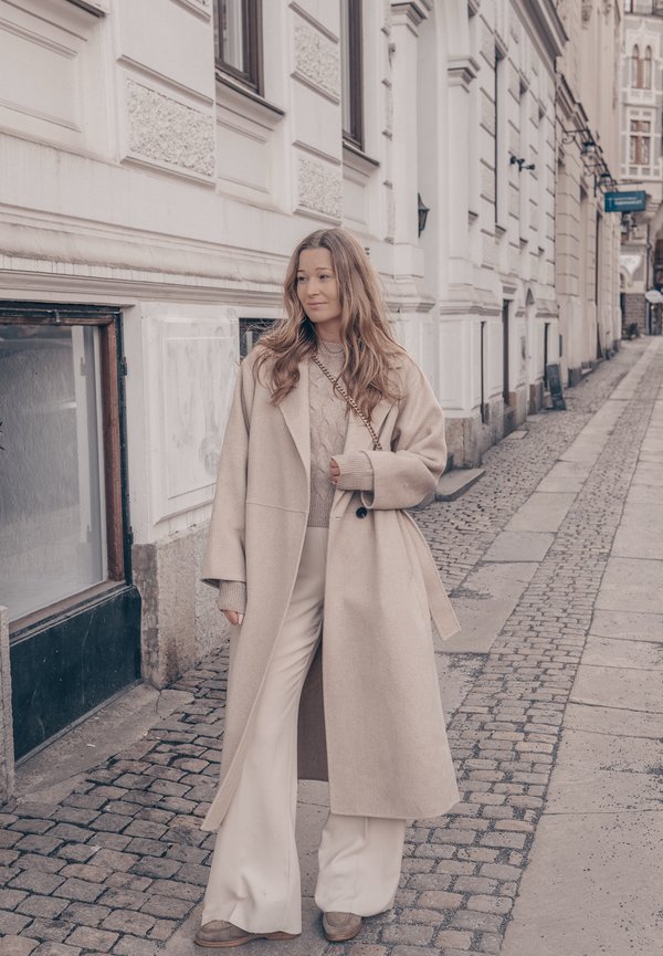 Femme en manteau beige long, pull en maille torsadée et pantalon large blanc, debout sur une rue pavée à côté d'un bâtiment blanc orné.