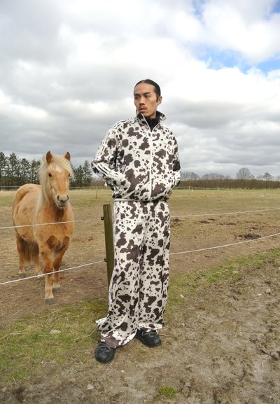 Chándal de patrón de vaca en blanco y negro con chaqueta de cremallera y pantalones sueltos, combinado con zapatos negros. Un pony de color marrón claro está cerca.