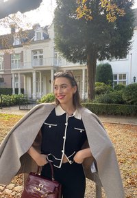 Μαύρος cropped μπλούζα με λευκή επένδυση και κουμπιά, συνδυασμένη με ένα ανοιχτό γκρι wrap. Καφέ τσάντα χειρός και γυαλιά ηλίου. Φθινοπωρινό φόντο με πράσινα φυτά.