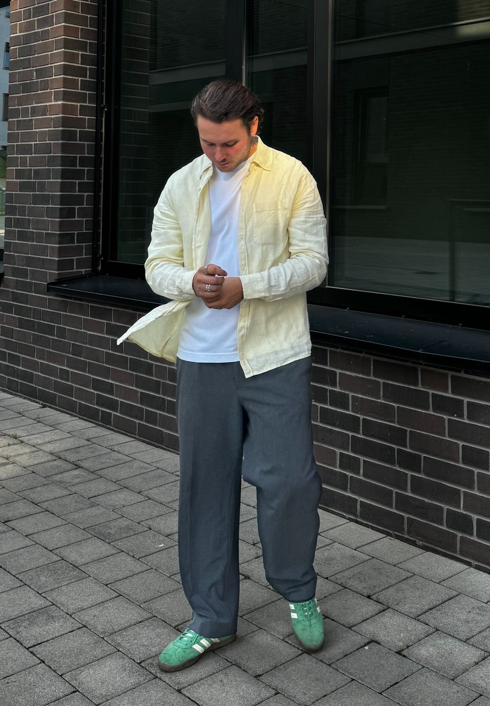 Világos sárga gombos ing, fehér póló, szürke, bőszárú nadrág és zöld sportcipő fehér csíkkal. Tégla burkolatú felületen áll.
