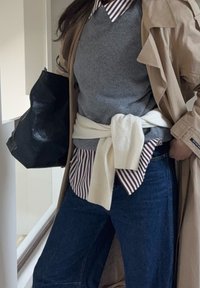 Pull gris superposé sur une chemise blanche à rayures bordeaux, assorti à un jean foncé. Un pull crème est noué autour de la taille, à côté d'un sac noir.