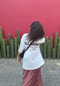 T-shirt oversized branca com o logótipo "ALLSAINTS" nas costas, saia longa vermelha estampada, de pé perto de cactos verdes contra uma parede rosa.