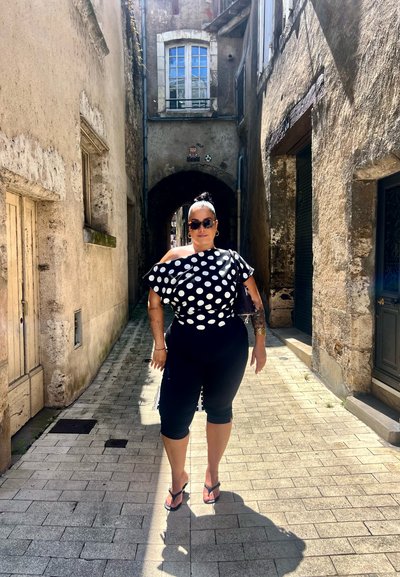 Top negro y blanco con lunares, sin mangas, combinado con pantalones negros recortados, de pie en una calle empedrada en un callejón estrecho.