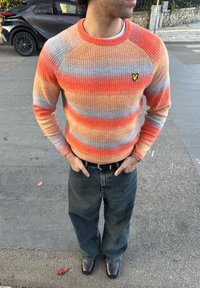 Pull à côtes multicolore avec rayures orange, bleu et crème ; col rond ; petit logo noir sur le côté gauche de la poitrine ; associé à un jean ample foncé.
