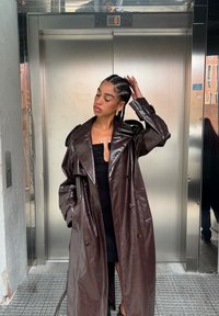 Brun trenchcoat i konstläder med bred krage, dubbelknäppt knappar och justerbara manschetter, bärs över en svart figurnära klänning.