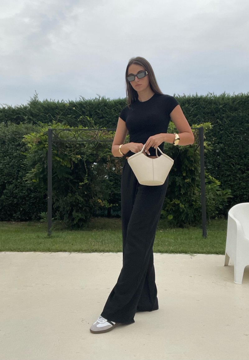 Top nero aderente, pantaloni neri a vita alta e larghi, borsa bianca con una forma strutturata, dettagli in argento sulle scarpe, ambiente esterno con vegetazione.