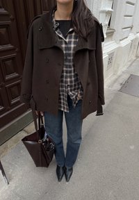 Manteau en laine brun croisé, chemise à carreaux, jean bleu, bottes noires à bout pointu, et sac fourre-tout en cuir bordeaux. Matériaux texturés, coupe décontractée.